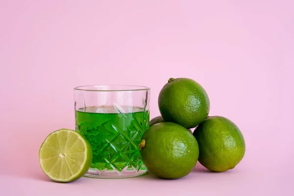 Green liquid in glass near fresh limes on pink background — Photo de stock