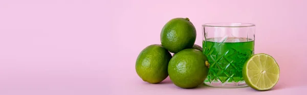 Green liquid in glass near fresh limes on pink background, banner — Stock Photo
