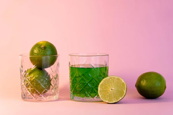 Green alcohol drink near faceted glass with fresh limes on pink background — Foto stock