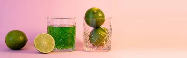 Grünes Alkoholgetränk neben facettiertem Glas mit frischen Limetten auf rosa Hintergrund, Banner — Stockfoto