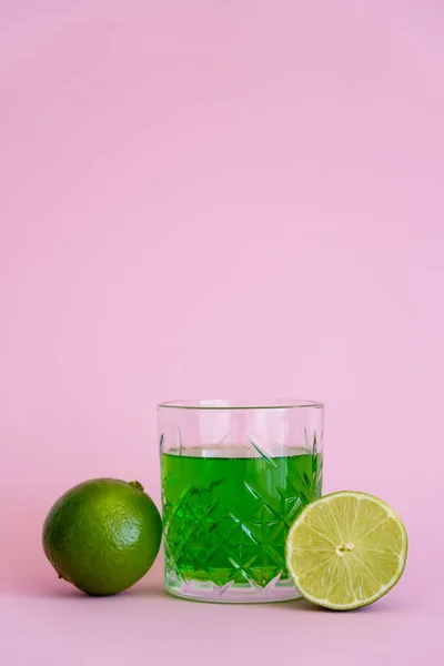Bebida alcohólica verde en vidrio facetado cerca de limas frescas sobre fondo rosa - foto de stock