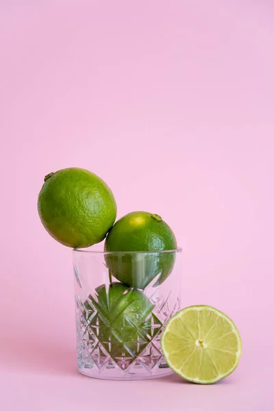 Grüne frische Zitrusfrüchte im facettierten Glas nahe der Hälfte der Limette auf rosa — Stockfoto