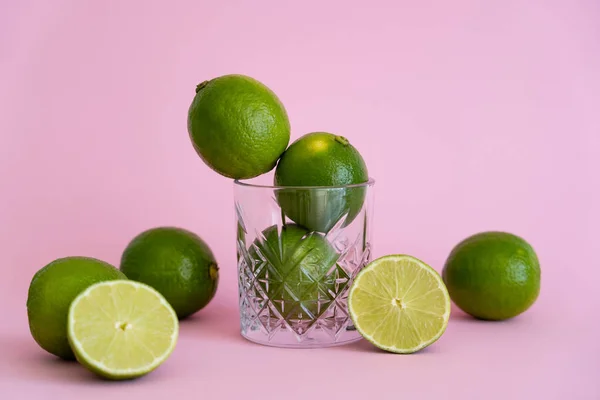 Green fresh citrus fruit in faceted glass near halves of limes on pink background — Stock Photo