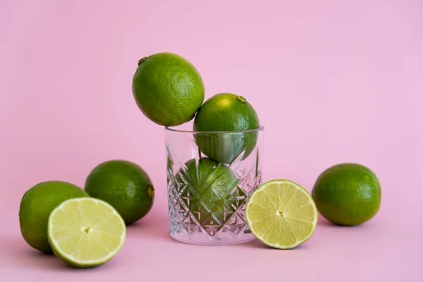 Cítricos frescos verdes en vidrio facetado cerca de mitades de limas sobre fondo rosa - foto de stock