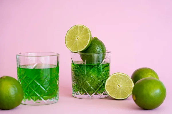 Green and fresh limes near faceted glasses with alcohol drink on pink — Photo de stock