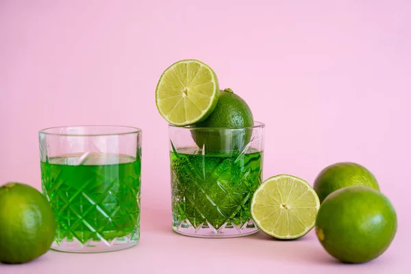 Green and fresh limes near faceted glasses with alcohol drink on pink — Stockfoto