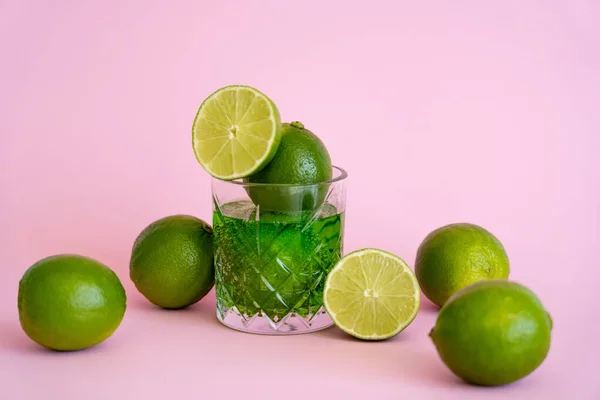 Grüne und frische Limetten im facettierten Glas mit Alkoholgetränk auf rosa — Stockfoto