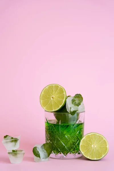 Green and fresh limes in faceted glass with sparkling alcohol drink near ice cubes with peppermint on pink - foto de stock
