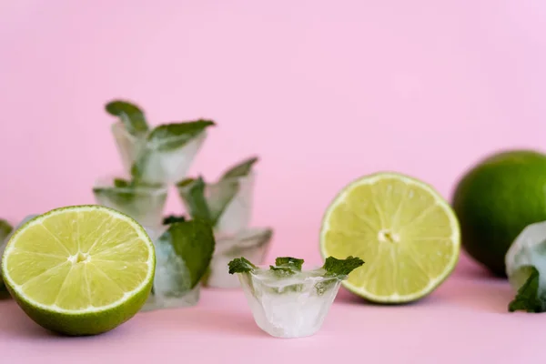 Green and fresh limes near blurred ice cubes with peppermint on pink — Stock Photo