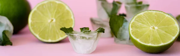 Green and fresh limes near blurred ice cubes with peppermint on pink, banner - foto de stock