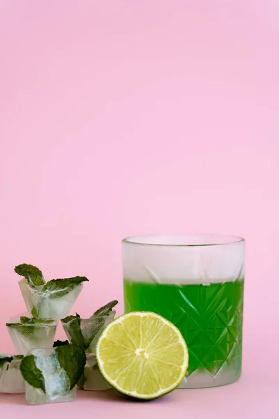 Green and fresh lime in faceted glass with alcohol drink near ice cubes with peppermint on pink — Stock Photo