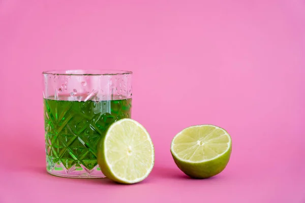 Green alcohol drink in faceted glass with water drops near limes on pink — Stock Photo