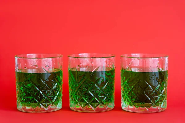 Boisson alcoolisée verte en trois verres à facettes avec gouttes d'eau sur rouge — Photo de stock