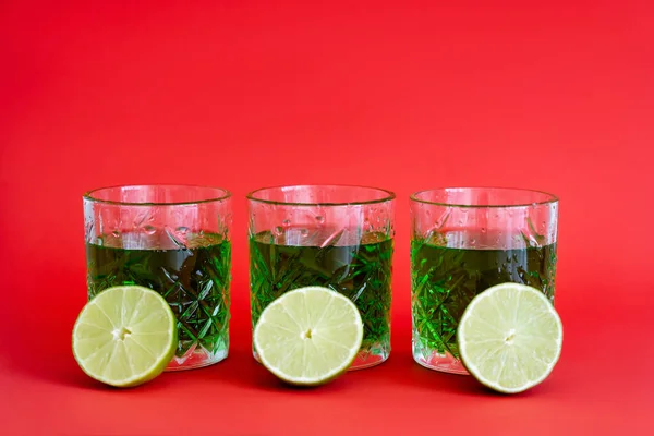 Green alcohol drink in three faceted glasses with water drops and halves of limes on red — Foto stock