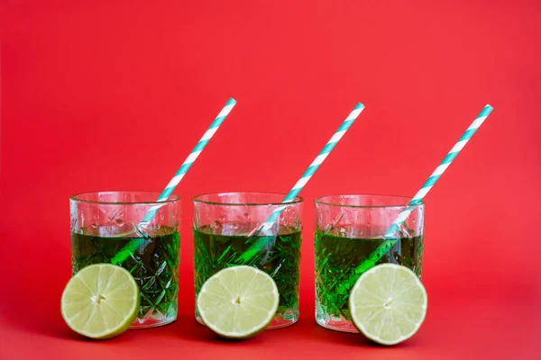 Bebida alcohólica verde en vasos facetados con pajitas y mitades de limas sobre rojo - foto de stock