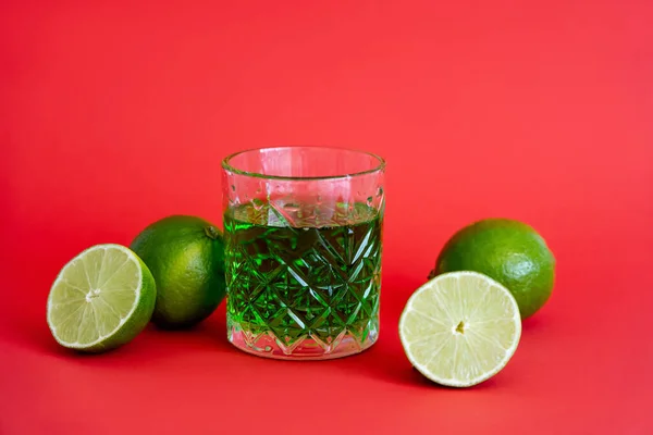 Green alcohol drink in faceted glass near sour limes on red — Stock Photo