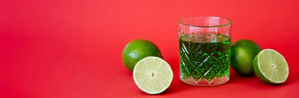 Green alcohol drink in faceted glass near sour limes on red, banner — Foto stock