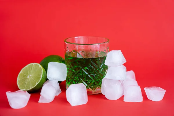 Glaçons surgelés près du verre avec mojito vert alcool et citrons verts sur rouge — Photo de stock