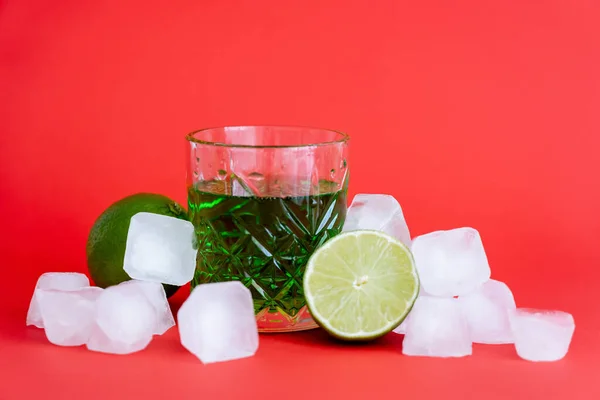 Frozen ice cubes near glass with alcohol green drink and limes on red — Foto stock