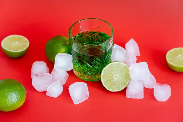 Cubitos de hielo congelados cerca del vidrio con mojito de alcohol y limas frescas sobre rojo - foto de stock