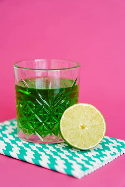 Glass of green alcohol drink on striped paper straws near sour lime on pink — Stock Photo