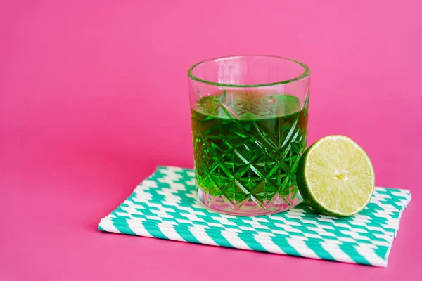 Glass of green alcohol drink on striped paper straws near sliced lime on pink — Stock Photo