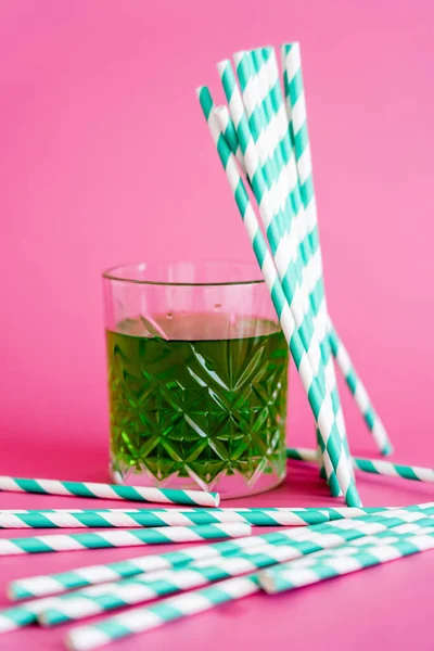 Faceted glass of green alcohol drink near striped paper straws on pink — Stock Photo