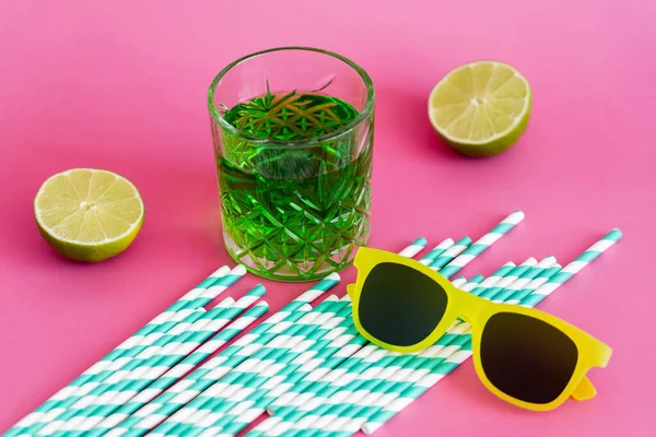 Lunettes de soleil et verre de boisson alcoolisée verte près de pailles et de citrons verts en papier rayé sur rose — Photo de stock