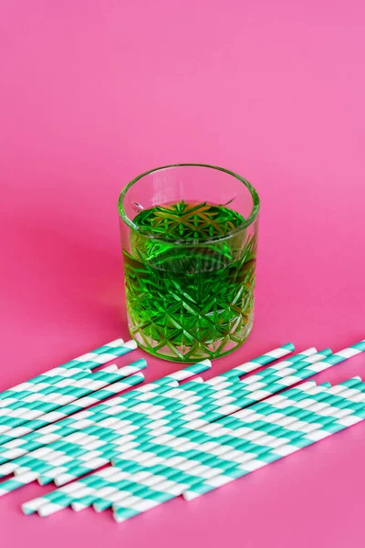 Pailles de papier rayé près du verre à facettes avec boisson verte sur rose — Photo de stock