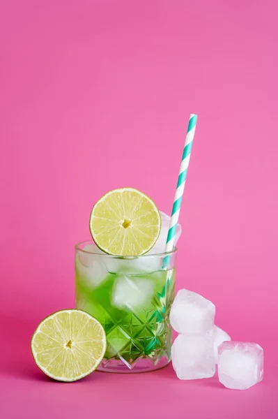 Frozen ice cubes in glass with green mojito drink with straw and limes on pink — Stock Photo