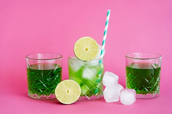 Frozen ice cubes in glass with mojito near green beverages and limes on pink — Stock Photo