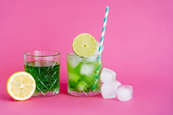 Ice cubes in glass with mojito near green beverage and limes on pink — Stockfoto