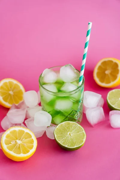 Frozen ice cubes in glass with mojito drink and straw near citrus fruits on pink — Stock Photo