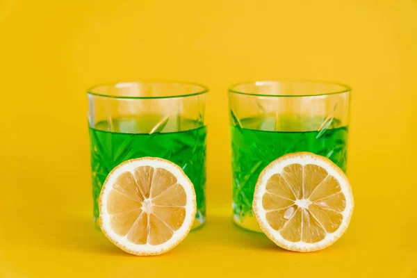 Bicchieri sfaccettati con bevanda alcolica verde e limoni affettati su giallo — Foto stock