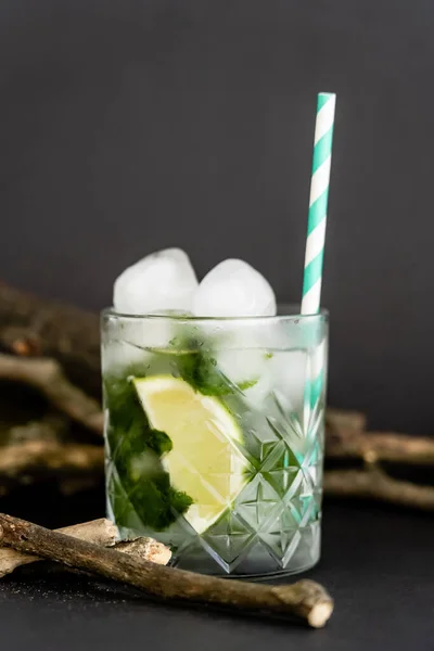 Faceted cold glass with ice cubes and sliced lime near wooden sticks on black - foto de stock