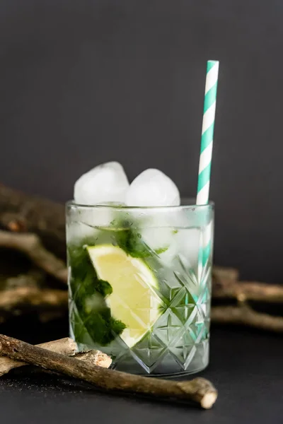 Vidrio frío facetado con cubitos de hielo y lima en rodajas cerca de palos de madera en negro - foto de stock