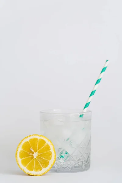 Faceted cold glass with ice cubes and paper straw near sliced lemon on white — Photo de stock