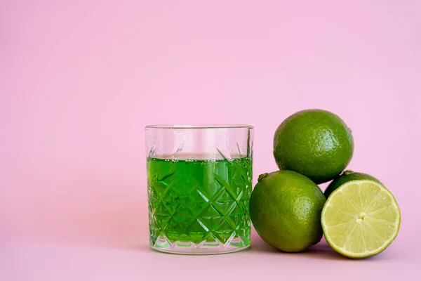 Grünalkoholgetränk im Glas in der Nähe von frischen und Bio-Limetten auf rosa — Stockfoto
