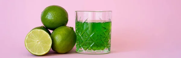 Grünes Alkoholgetränk im Glas in der Nähe von frischen und biologischen Limetten auf rosa, Banner — Stockfoto