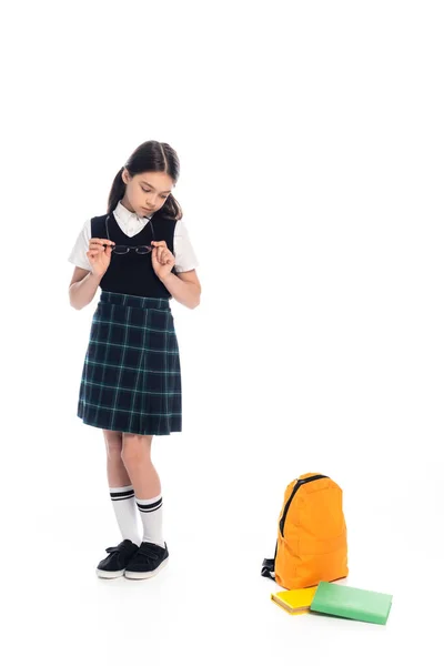 Задумчивый школьник держит очки возле книг и рюкзака на белом фоне — стоковое фото