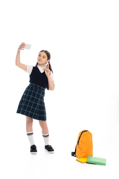 Школьник показывает знак мира, делая селфи на смартфоне возле книг и рюкзака на белом фоне — стоковое фото
