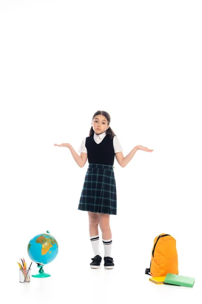 Niño confundido mostrando gesto de encogimiento de hombros cerca del globo y libros sobre fondo blanco - foto de stock