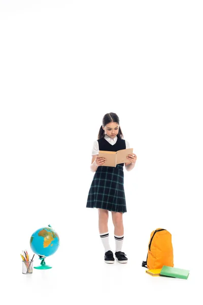Full length of schoolchild reading book near globe and color pencils on white background — Stock Photo