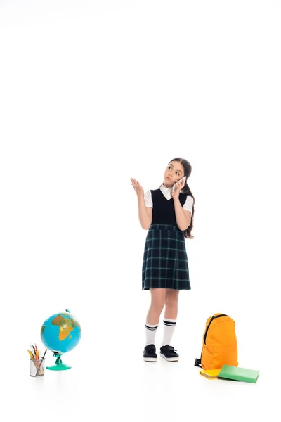 Pupil talking on smartphone and pointing with hand near globe and books on white background — Stock Photo