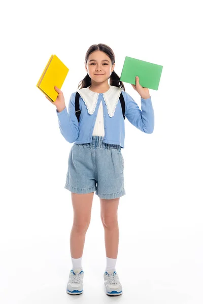 Écolière souriante en short denim tenant des livres sur fond blanc — Photo de stock