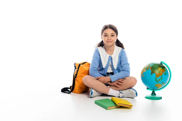 Lächelnder Schüler, der neben Büchern und Globus auf weißem Hintergrund sitzt — Stockfoto