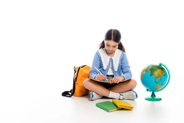 Preteen schoolkid writing on notebook near backpack and globe on white background — Stock Photo