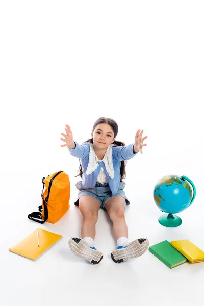 Vista de ángulo alto de la colegiala extendiendo las manos a la cámara cerca del globo y libros sobre fondo blanco - foto de stock