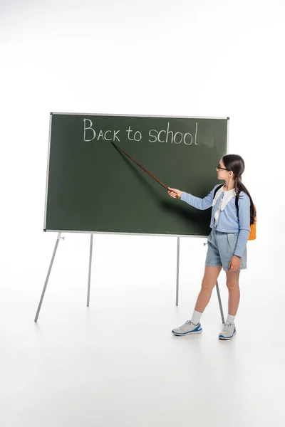 Vue latérale de l'écolier tenant pointeur près de retour à l'école lettrage sur tableau noir sur fond blanc — Photo de stock