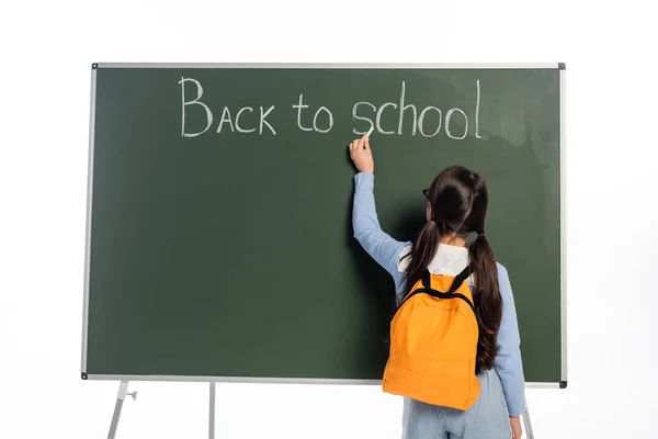 Rückansicht von Schulmädchen mit Rucksack, die Schulschriftzug auf Kreidetafel isoliert auf weiß schreibt — Stockfoto